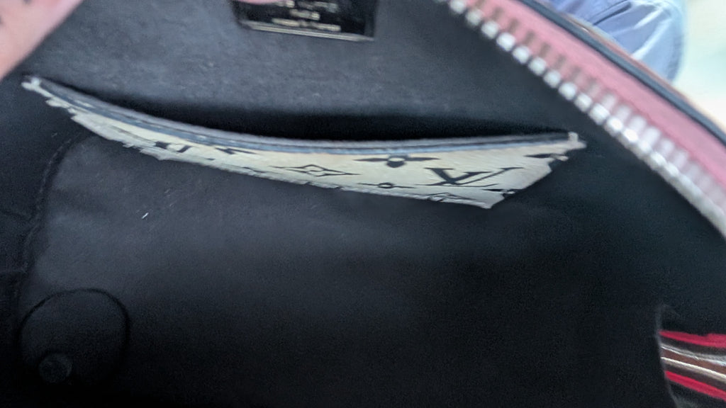 Close-up of the interior of a black handbag, showing a patterned paper tag attached inside a zippered pocket and a pink zipper edge. Pre-Owned LOUIS VUITTON ALMA BB – ROSE BLUSH MONOGRAM VERNIS MIROIR
