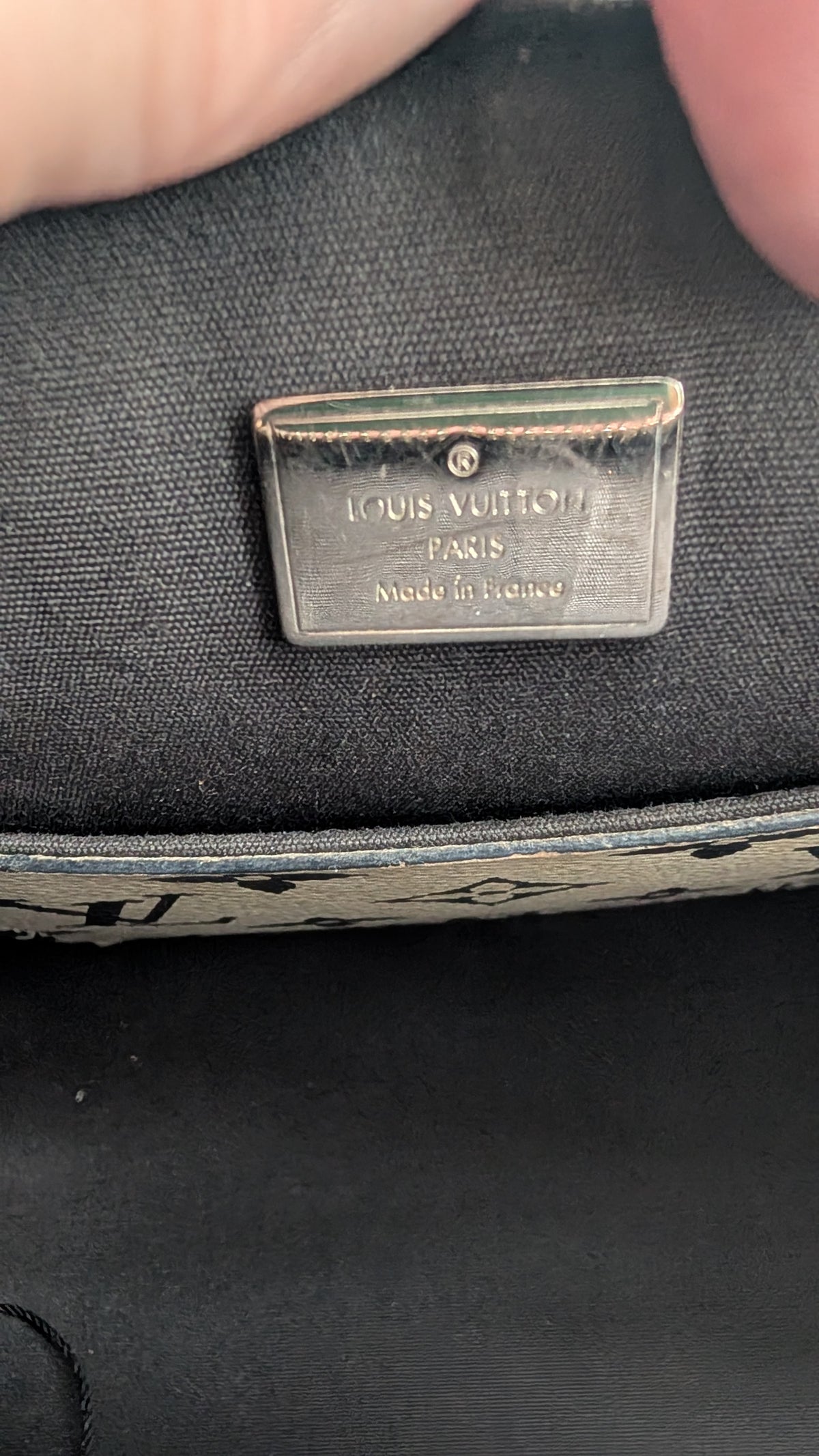 Close-up of a Louis Vuitton metal brand plaque on a black fabric surface, with fingers holding the item at the top edge. Pre-Owned LOUIS VUITTON ALMA BB – ROSE BLUSH MONOGRAM VERNIS MIROIR