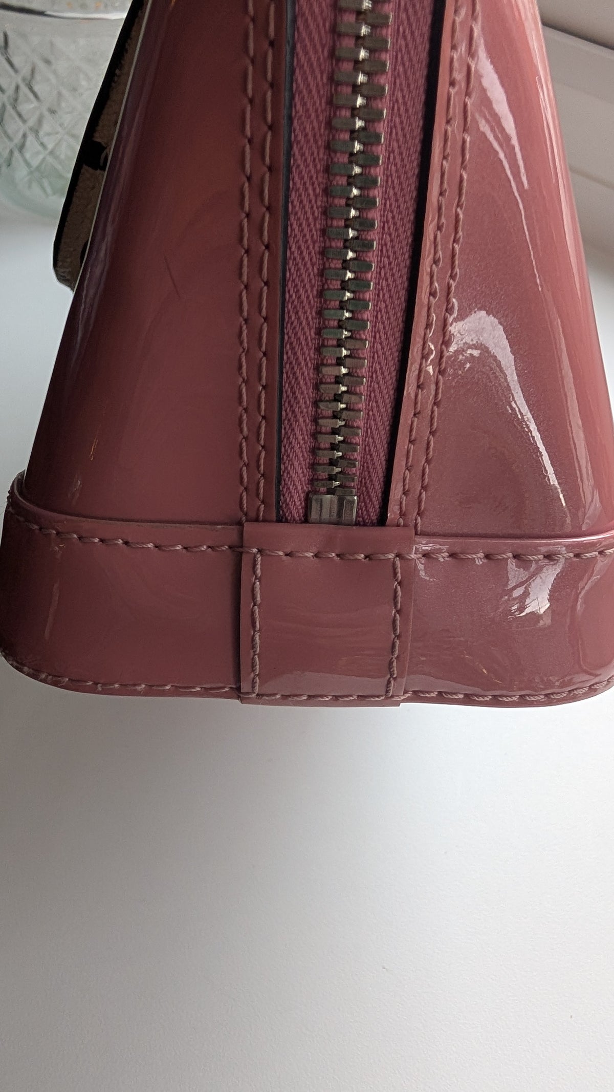 Close-up of a glossy patent leather handbag in dusty rose pink, showing a silver zipper and stitched details on a white surface. Pre-Owned LOUIS VUITTON ALMA BB – ROSE BLUSH MONOGRAM VERNIS MIROIR