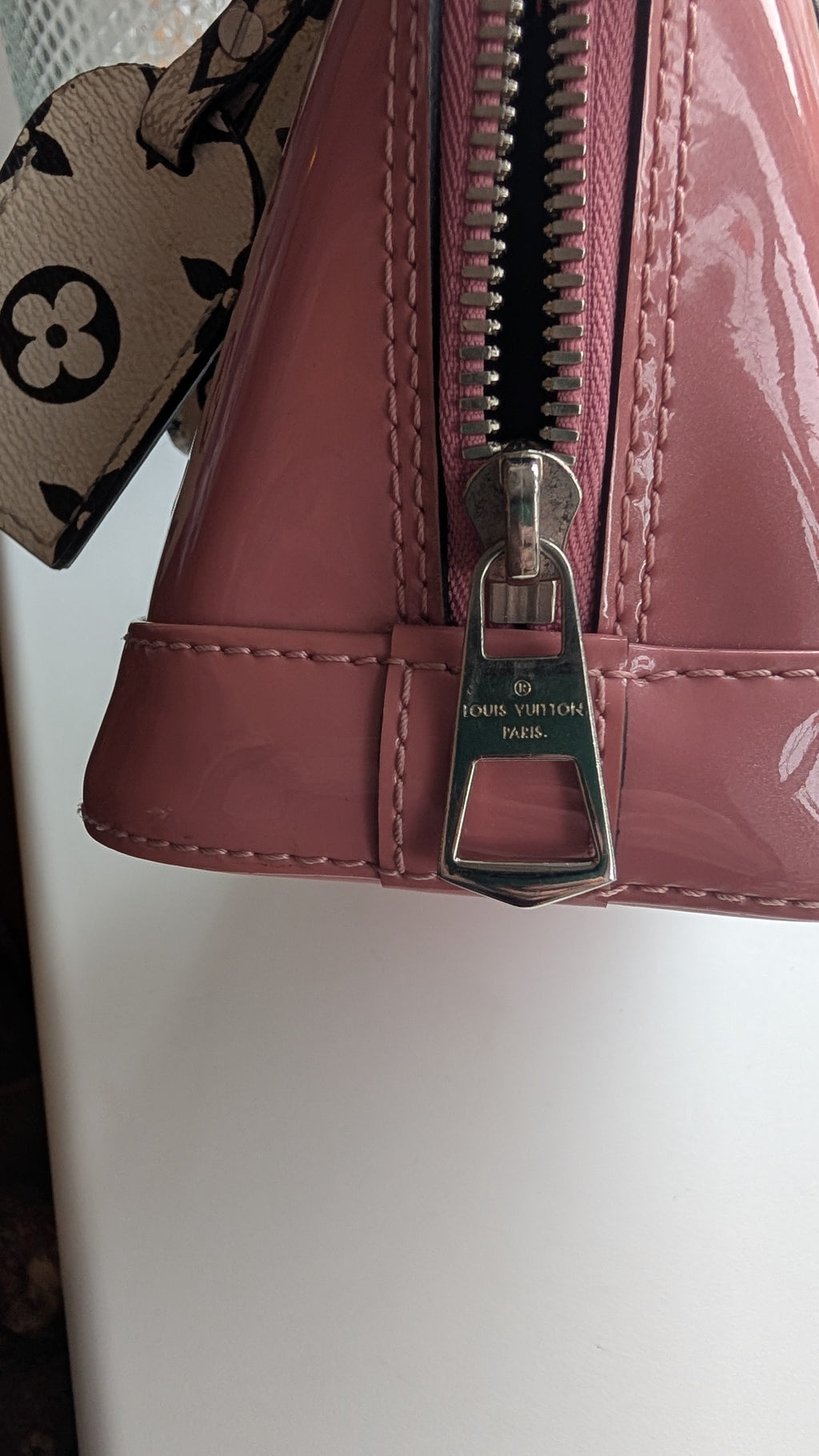 Close-up of a Louis Vuitton handbag in glossy pink patent leather, showing a silver-tone metal zipper pull engraved with 'LOUIS VUITTON PARIS'.Pre-Owned LOUIS VUITTON ALMA BB – ROSE BLUSH MONOGRAM VERNIS MIROIR