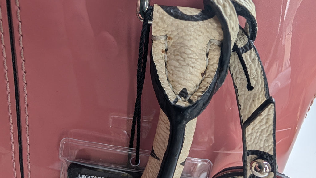 A close-up of a pink handbag showing a worn beige and black textured strap attached with a metal ring, alongside a clear plastic protective cover and a black hangtag.Pre-Owned LOUIS VUITTON ALMA BB – ROSE BLUSH MONOGRAM VERNIS MIROIR