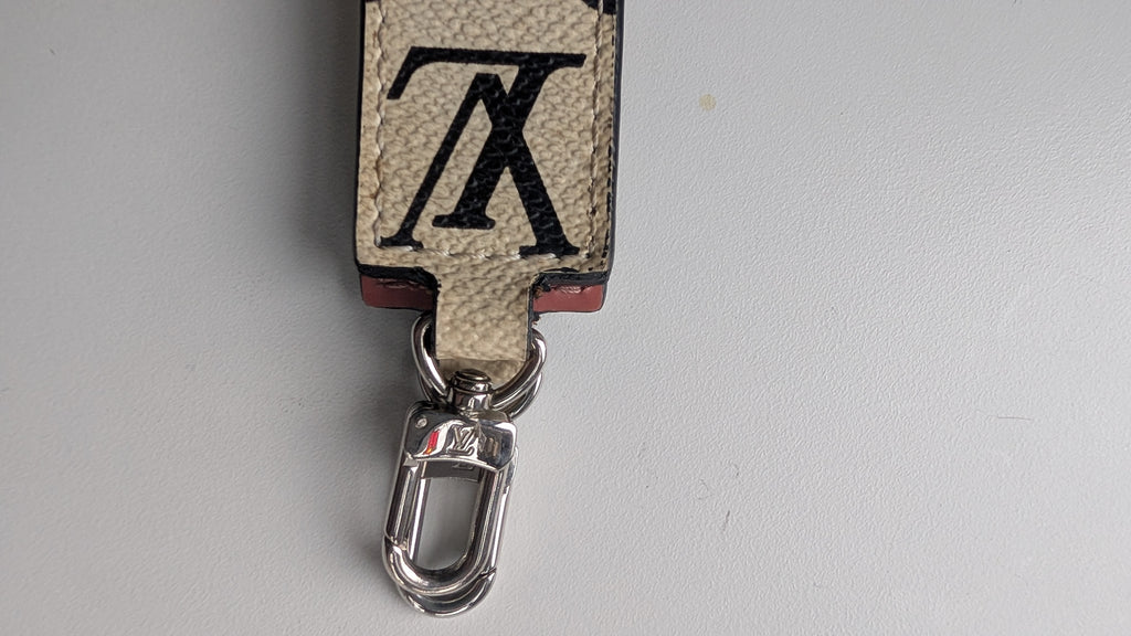 A close-up of a Louis Vuitton keychain featuring the brand's signature beige and black monogram canvas, a small red leather accent, and a silver metal lobster clasp, set against a plain light gray background.Pre-Owned LOUIS VUITTON ALMA BB – ROSE BLUSH MONOGRAM VERNIS MIROIR