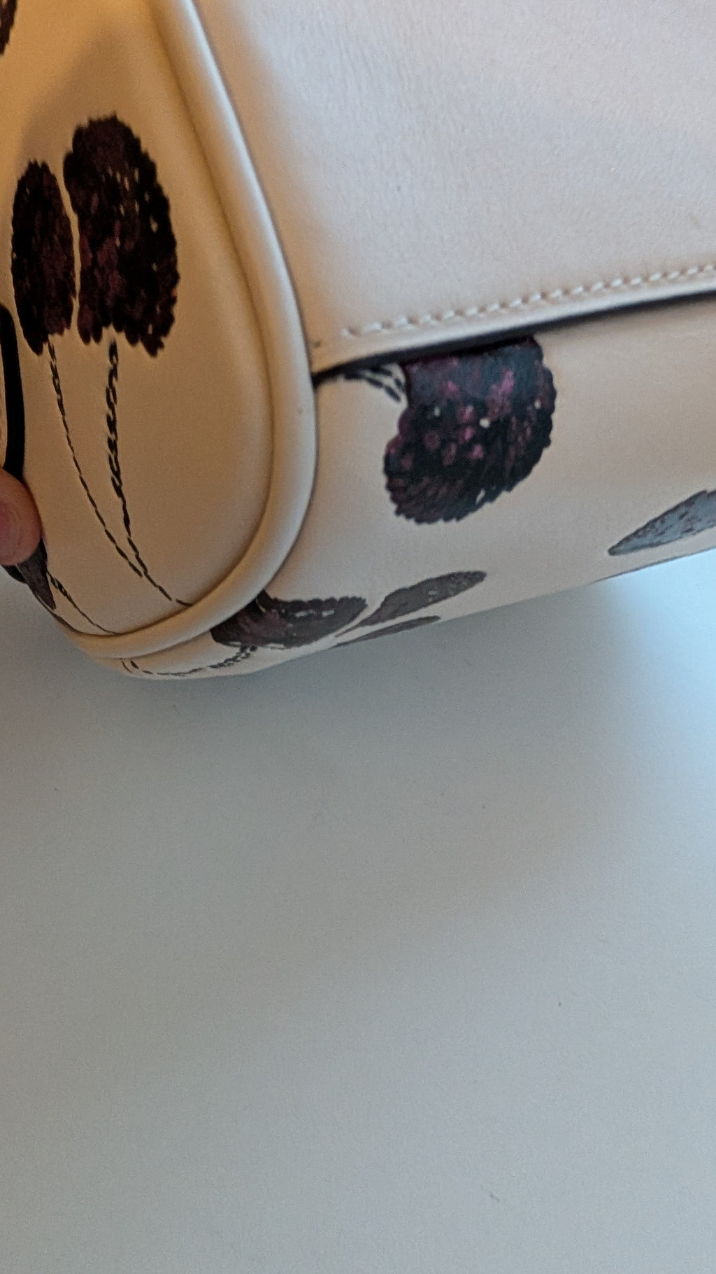 Close-up of a cream-colored handbag with a dark cherry pattern, showing the corner and a hand holding it to demonstrate scale. Pre-Owned Coach Rowan Cherry Print Leather Crossbody / Mini Satchel – Chalk Multi