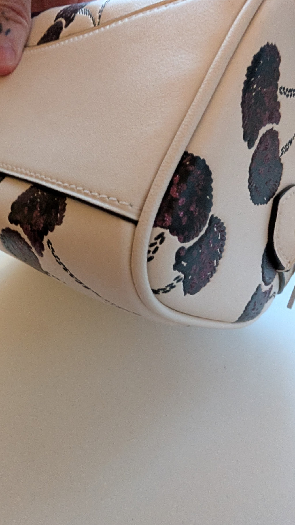 Close-up of a cream-colored handbag with a black cherry pattern on the side panel, held by a hand at the top left corner. Pre-Owned Coach Rowan Cherry Print Leather Crossbody / Mini Satchel – Chalk Multi