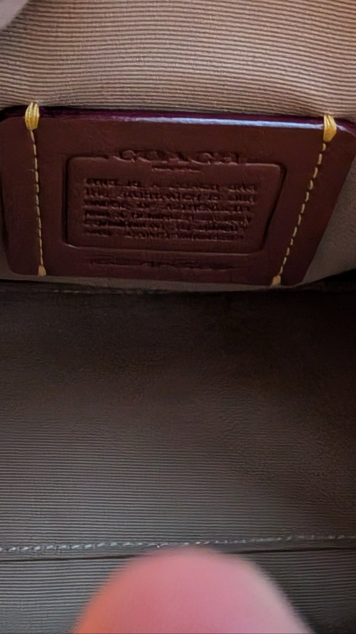 Close-up of the inside of a Coach handbag showing a brown leather brand patch, beige interior lining, and a hand with tattoos holding the bag open. Pre-Owned Coach Rowan Cherry Print Leather Crossbody / Mini Satchel – Chalk Multi