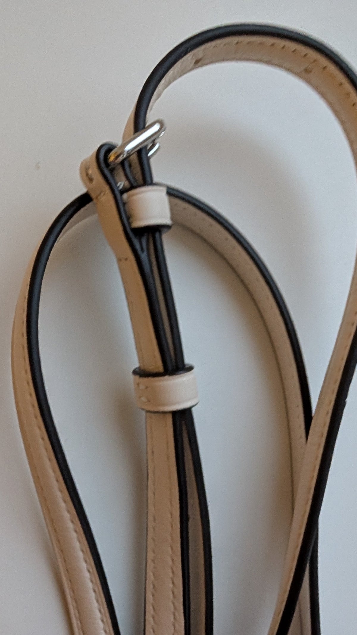 A close-up of a hand holding a tan leather strap with black edging and silver metal hardware against a plain light  Pre-Owned Coach Rowan Cherry Print Leather Crossbody / Mini Satchel – Chalk Multibackground.