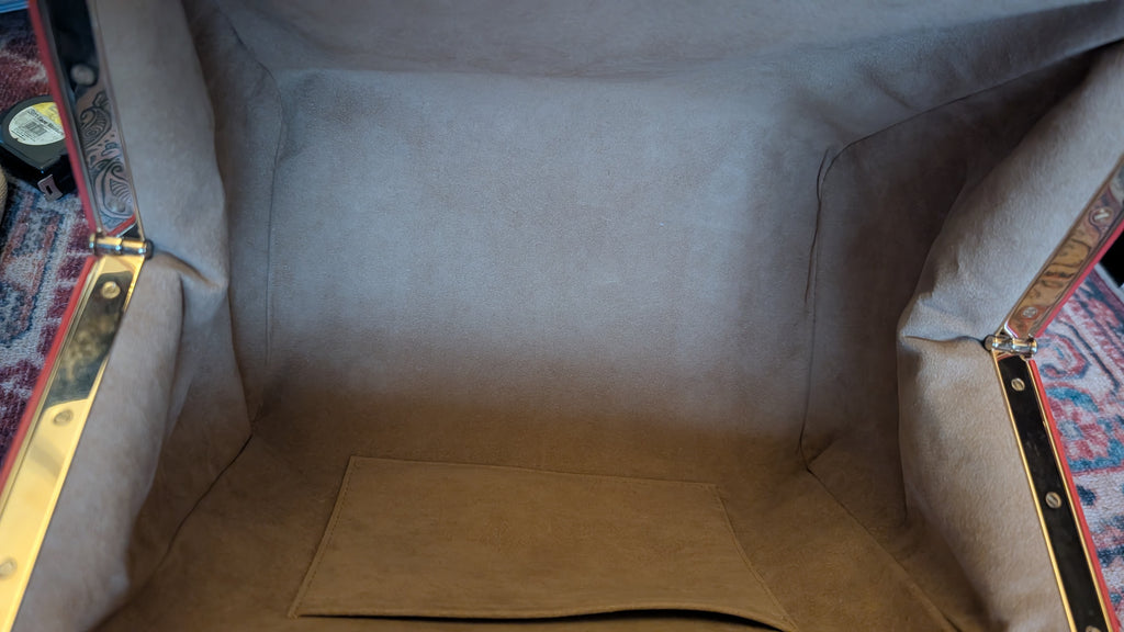 Close-up view inside a vintage upholstered trunk or storage compartment, showing beige fabric lining, brass hardware, and patterned rugs at the sides. Pre-Owned Louis Vuitton x Richard Prince Limited Edition Monogram Aquarelle Watercolor Frame Speedy 30 – 2008