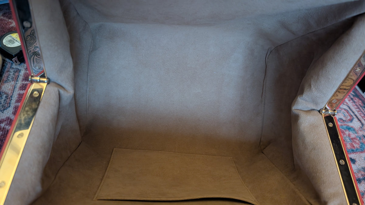 Close-up view inside a vintage upholstered trunk or storage compartment, showing beige fabric lining, brass hardware, and patterned rugs at the sides. Pre-Owned Louis Vuitton x Richard Prince Limited Edition Monogram Aquarelle Watercolor Frame Speedy 30 – 2008
