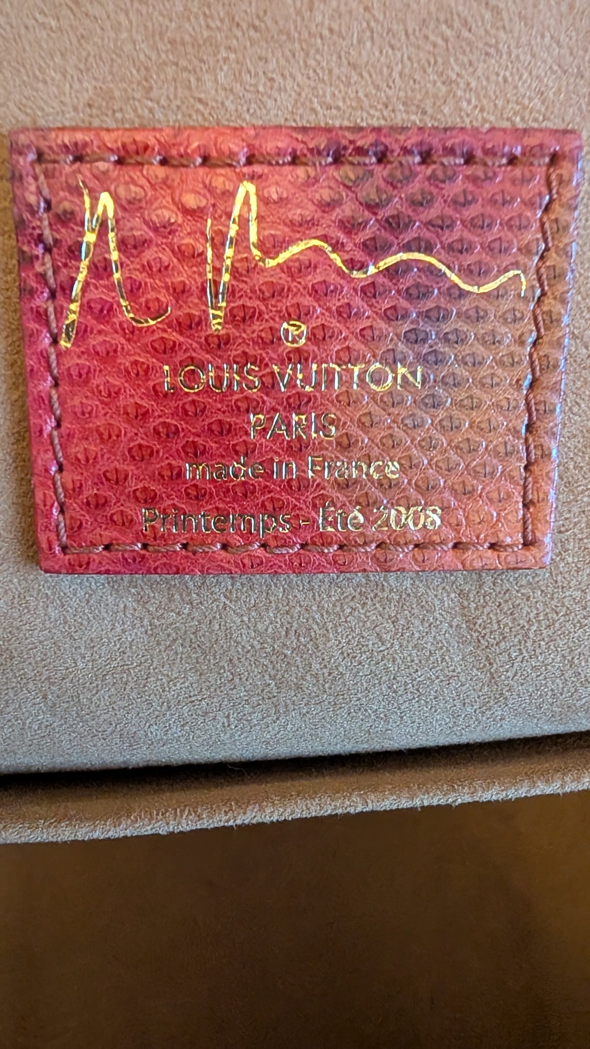 A close-up of a Louis Vuitton leather patch with gold foil stamping on a textured beige surface. The patch is pinkish-red with a subtle iridescent sheen and features the brand's signature zig-zag line logo. Pre-Owned Louis Vuitton x Richard Prince Limited Edition Monogram Aquarelle Watercolor Frame Speedy 30 – 2008