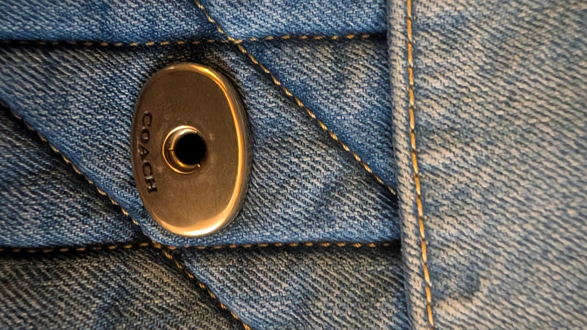 Close-up of a blue denim garment with a round gold-tone metal snap button engraved with the brand name COACH. Pre-Owned Coach Quilted Tabby Shoulder Bag 26 – Blue Denim