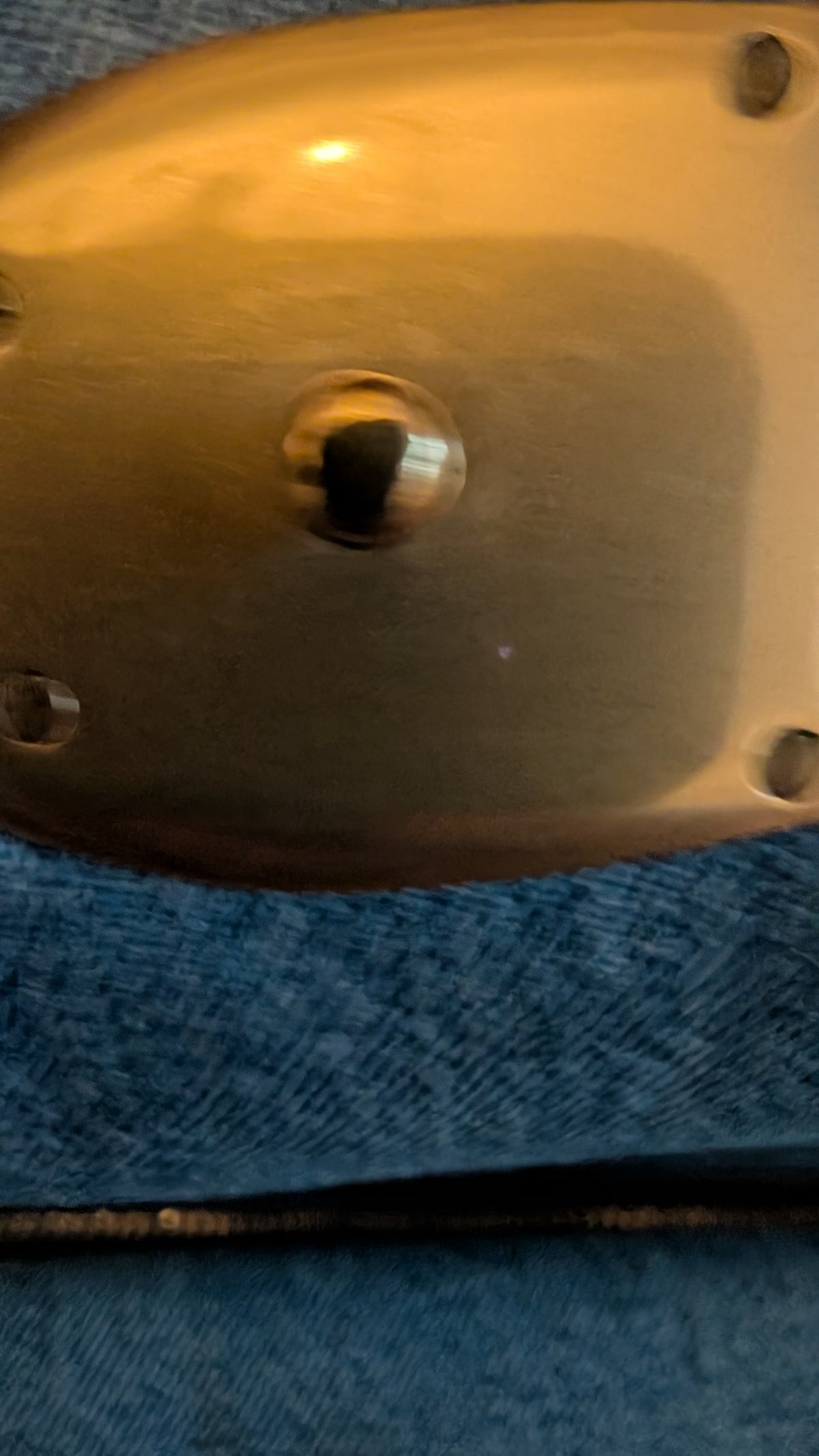 A close-up of a metallic gold-colored oval-shaped component with a central raised circular feature, resting on a textured blue fabric background. Pre-Owned Coach Quilted Tabby Shoulder Bag 26 – Blue Denim