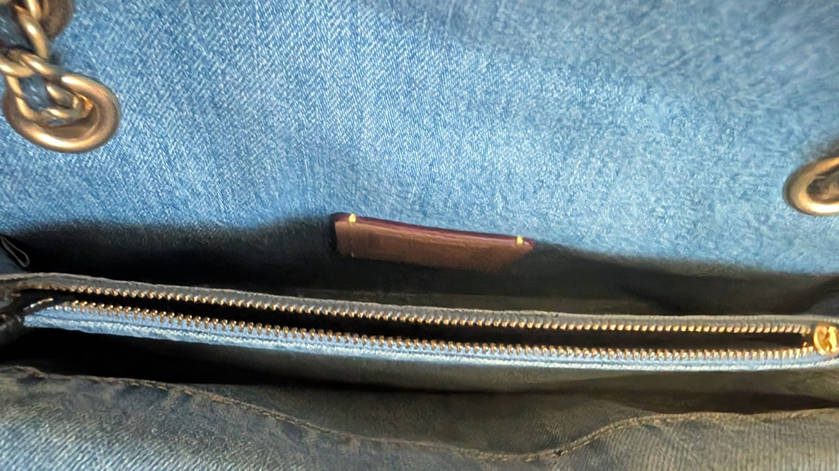 Close-up of the interior of a blue denim handbag, showing a gold zipper, a small brown leather brand tag, and part of a gold chain attachment. Pre-Owned Coach Quilted Tabby Shoulder Bag 26 – Blue Denim