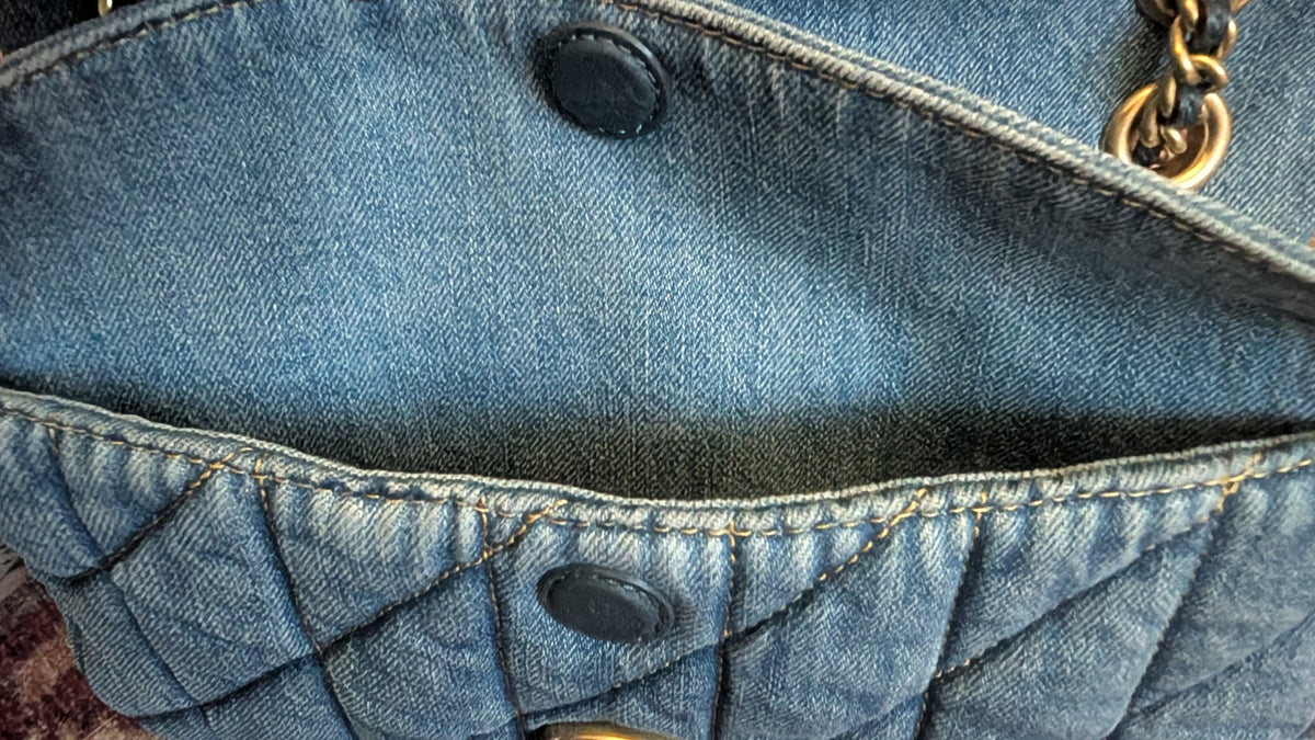 Close-up detail of a blue denim bag showing quilted stitching, dark snap buttons, and a gold-tone chain link. Pre-Owned Coach Quilted Tabby Shoulder Bag 26 – Blue Denim