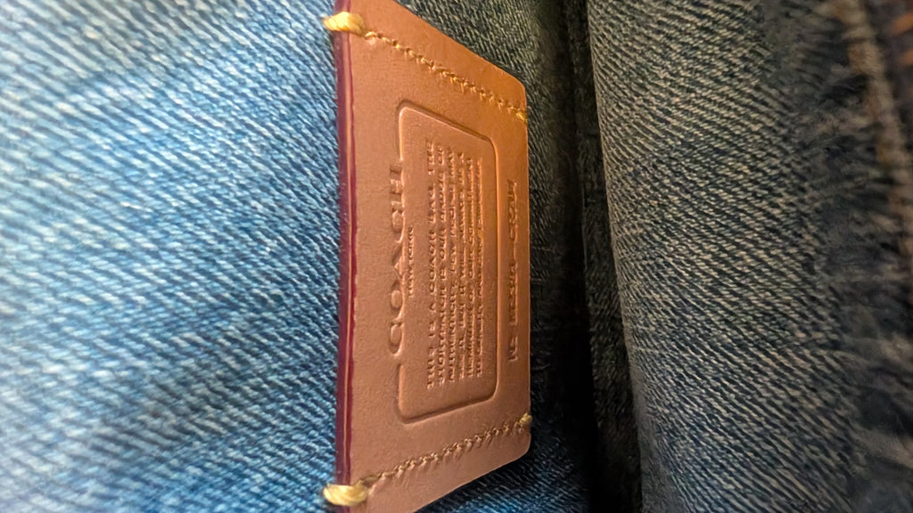A close-up of a tan leather Coach brand tag sewn onto blue denim fabric. Pre-Owned Coach Quilted Tabby Shoulder Bag 26 – Blue Denim