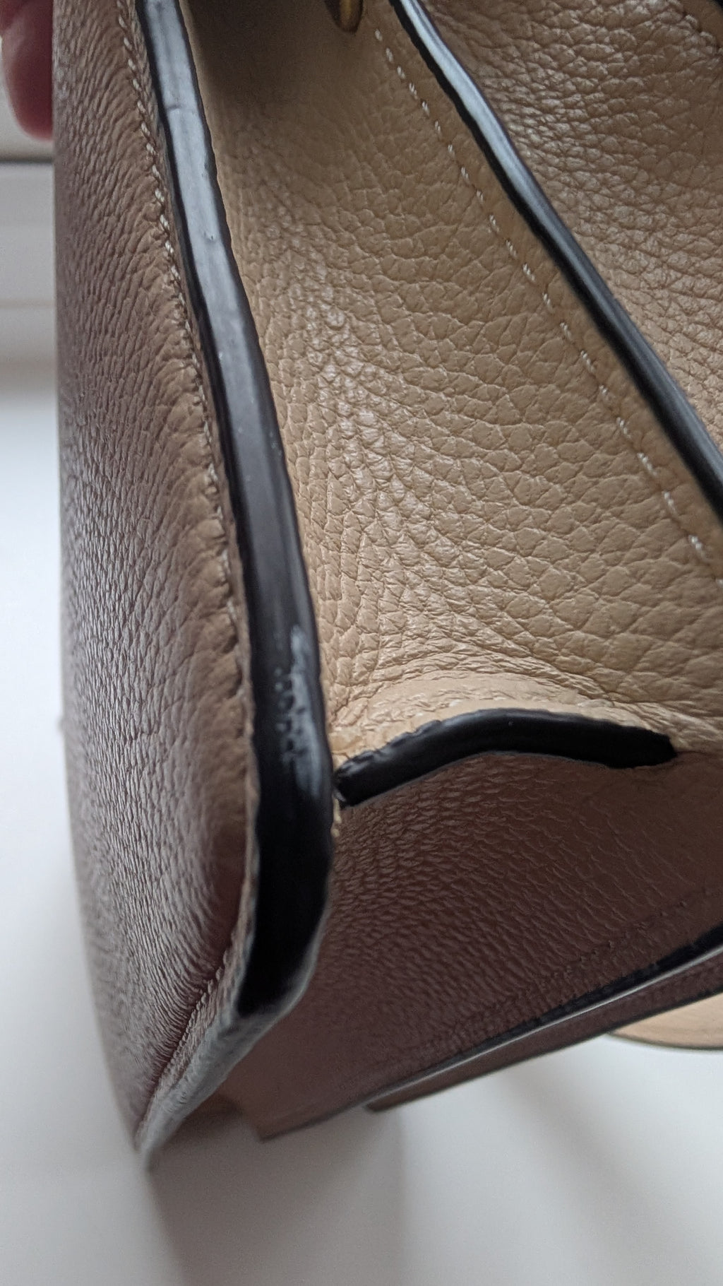 Close-up of a beige pebbled leather handbag corner showing the interior side with black trim and a brown base, photographed on a white surface. Pre-Owned Coach Tabby Braided Leather Shoulder Bag – Beige