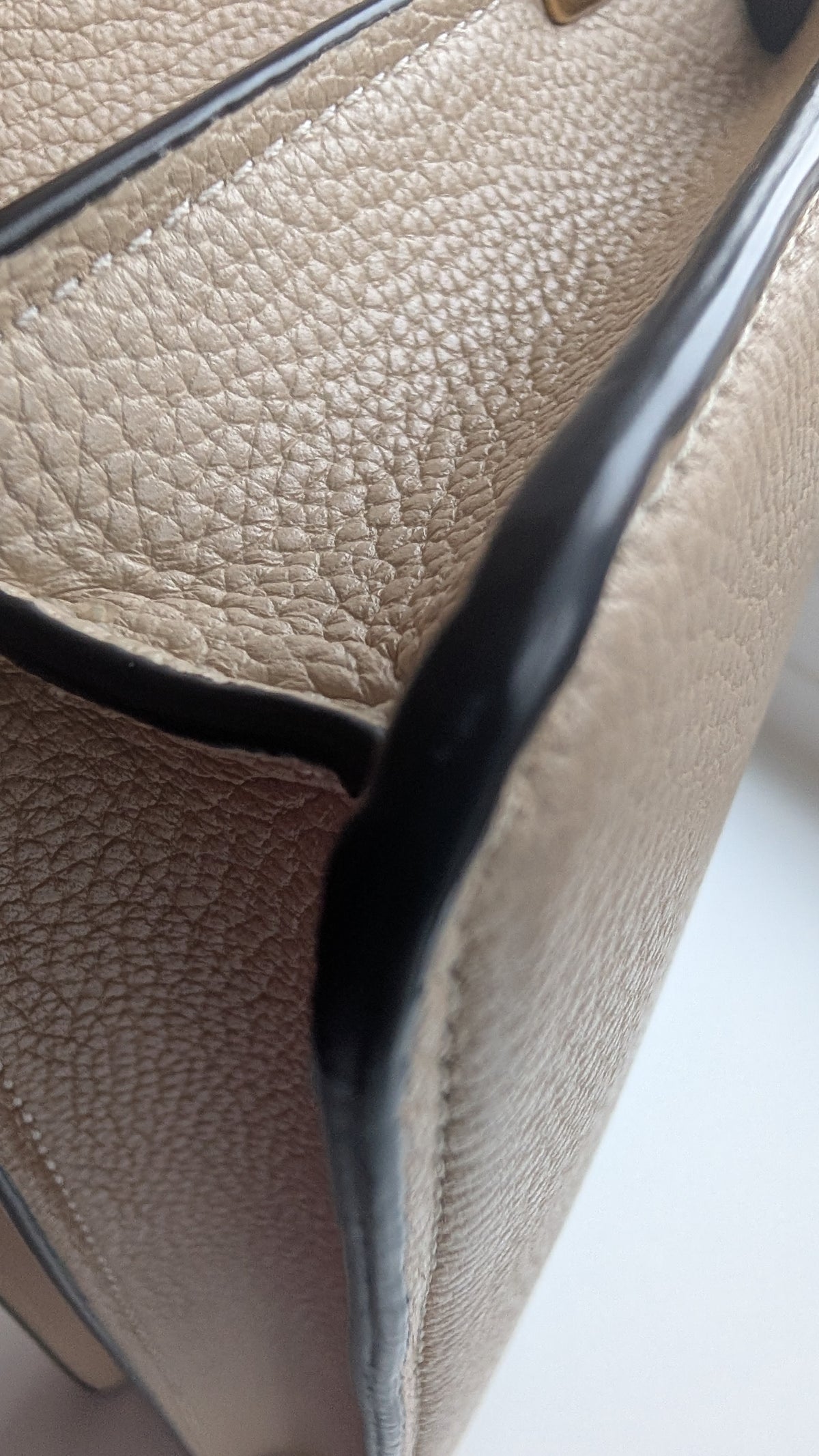 Close-up of a beige pebbled leather handbag corner with black trim and a small gold hardware accent. Pre-Owned Coach Tabby Braided Leather Shoulder Bag – Beige