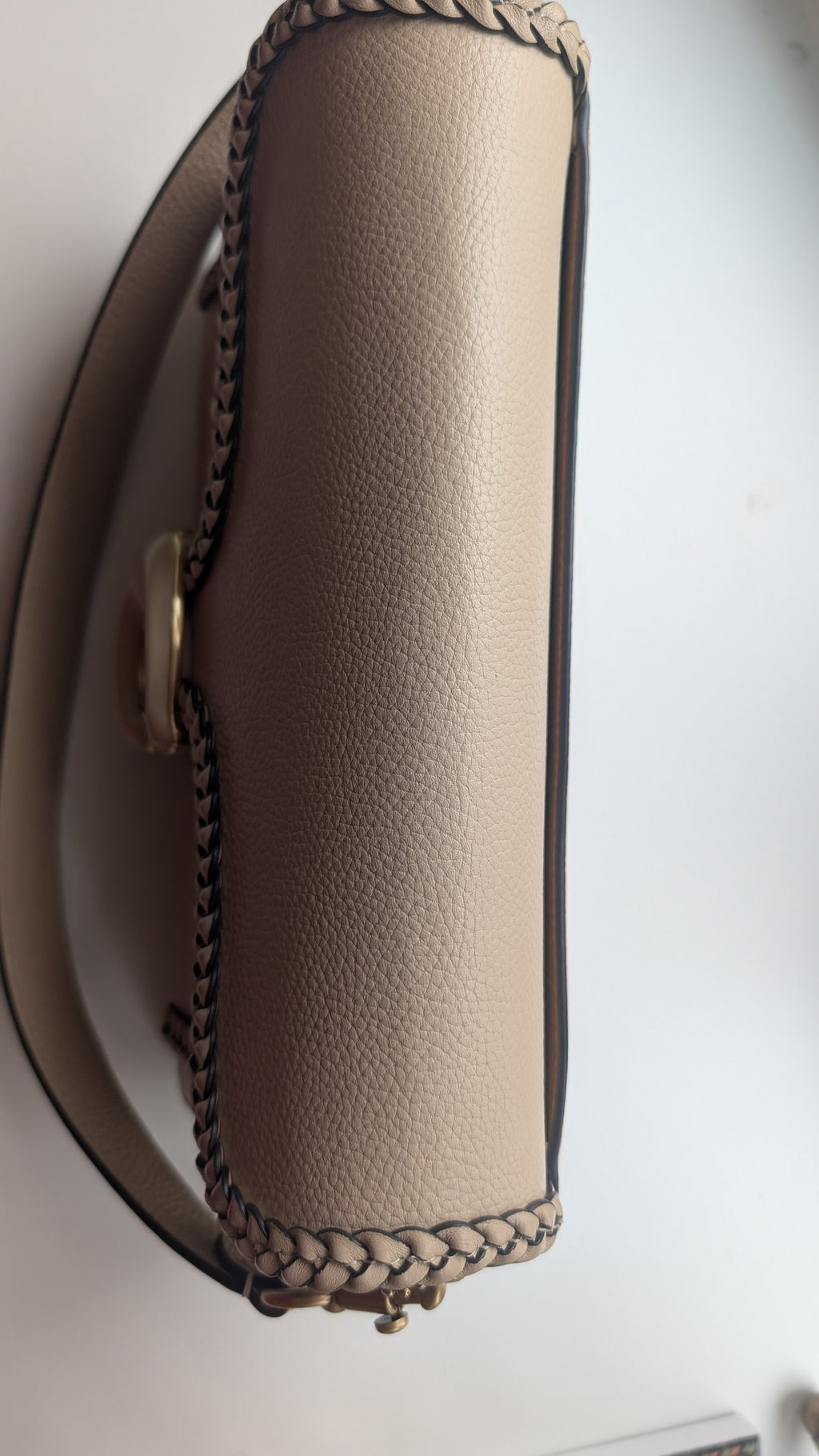 A close-up side view of a beige pebbled leather handbag with dark contrast stitching and a gold-tone clasp, photographed against a plain light background. Pre-Owned Coach Tabby Braided Leather Shoulder Bag – Beige