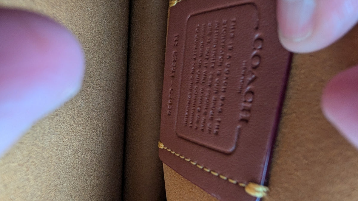 A close-up of a brown leather Coach brand tag being held by fingers against a tan textured background. Pre-Owned Coach Tabby Braided Leather Shoulder Bag – Beige