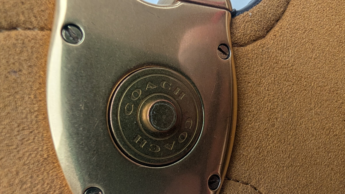 Close-up of a Coach logo button on a metallic bronze-toned leather accessory, set against a tan leather background. Pre-Owned Coach Tabby Braided Leather Shoulder Bag – Beige