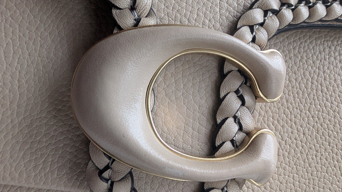 Close-up of a large metallic C-shaped hardware detail on a light beige textured leather bag, with braided strap details visible around it. Pre-Owned Coach Tabby Braided Leather Shoulder Bag – Beige