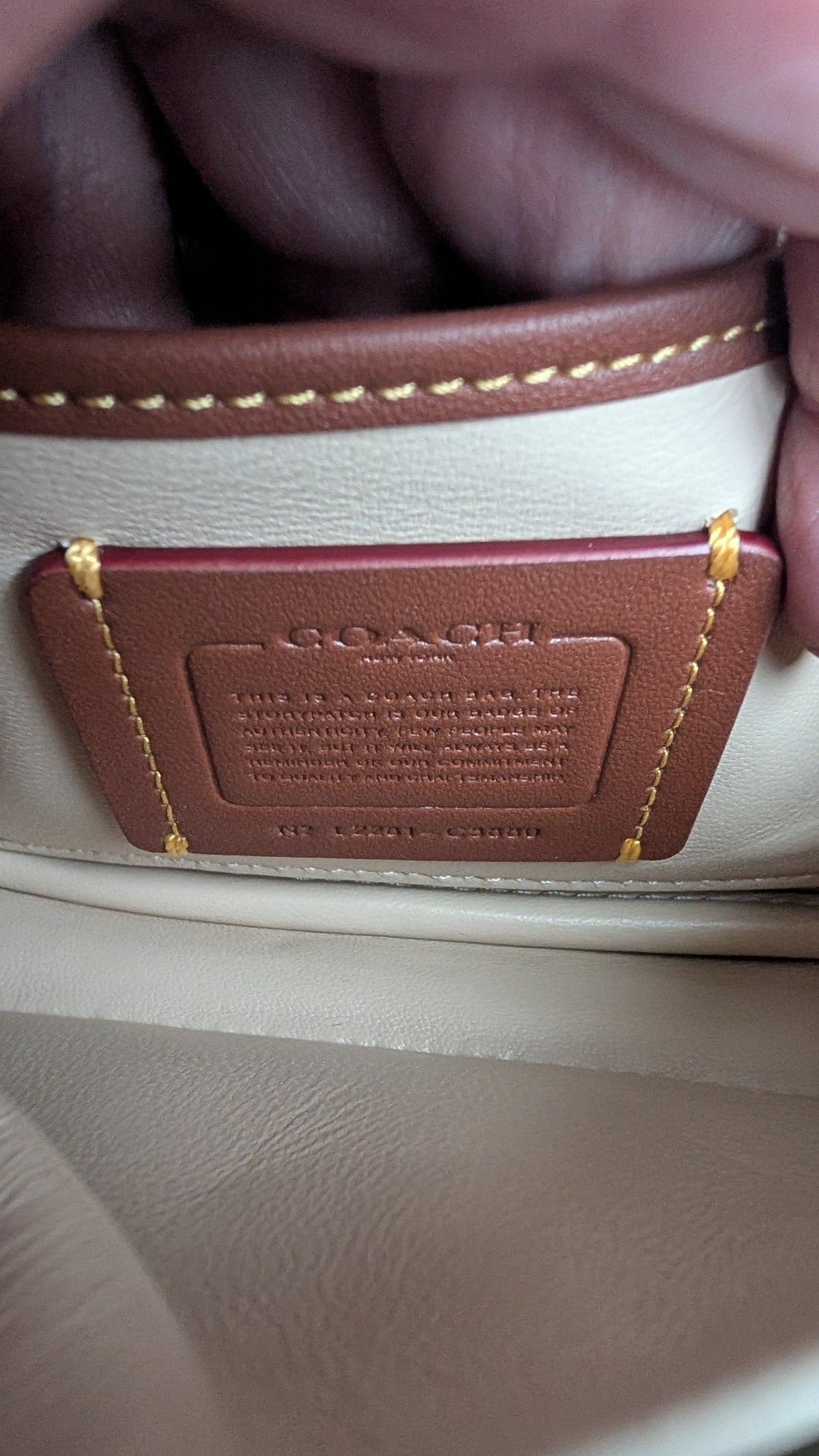 Close-up of a brown leather interior label with gold stitching, showing the embossed brand name Coach, held against a cream-colored leather background. Pre-Owned Coach Pillow Tabby Shoulder Bag – Cream/Ivory Leather