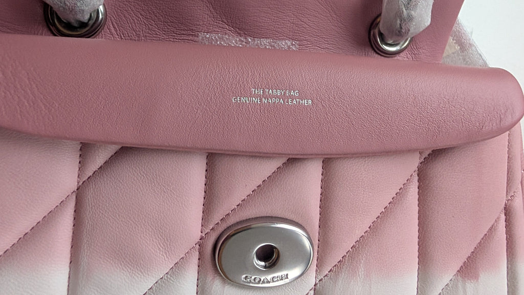 Close-up of a pink quilted leather handbag showing the flap, silver-tone hardware, and a small embossed brand mark on the leather tab. Pre-Owned Coach Tabby 20 Quilted Nappa Leather Shoulder Bag – Pink & White Ombré