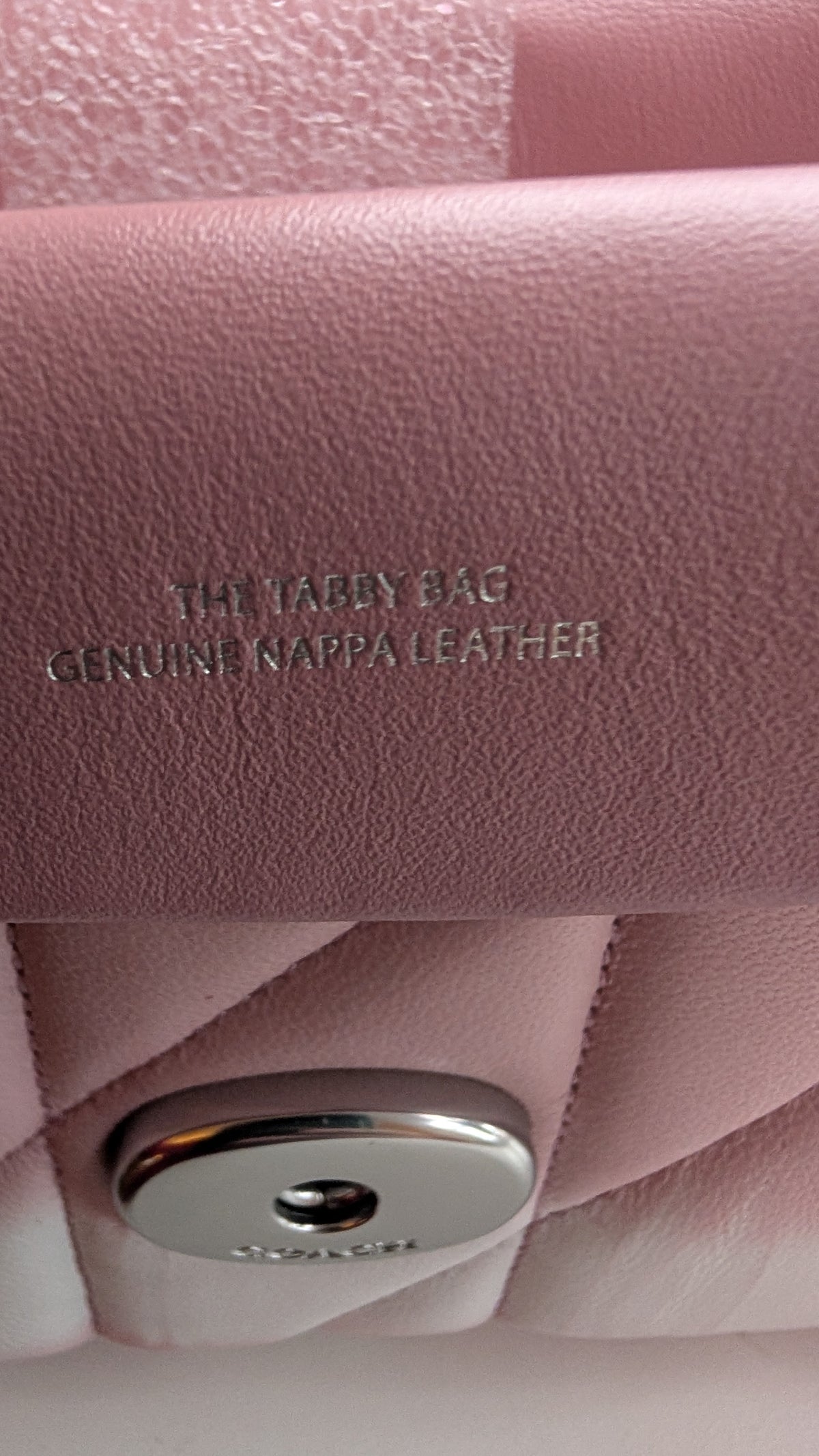 Close-up of a pink leather handbag flap with embossed text reading 'THE TAbby BAG' and 'GENUINE nAPPA LEATHER', plus a silver-tone metal snap closure. Pre-Owned Coach Tabby 20 Quilted Nappa Leather Shoulder Bag – Pink & White Ombré