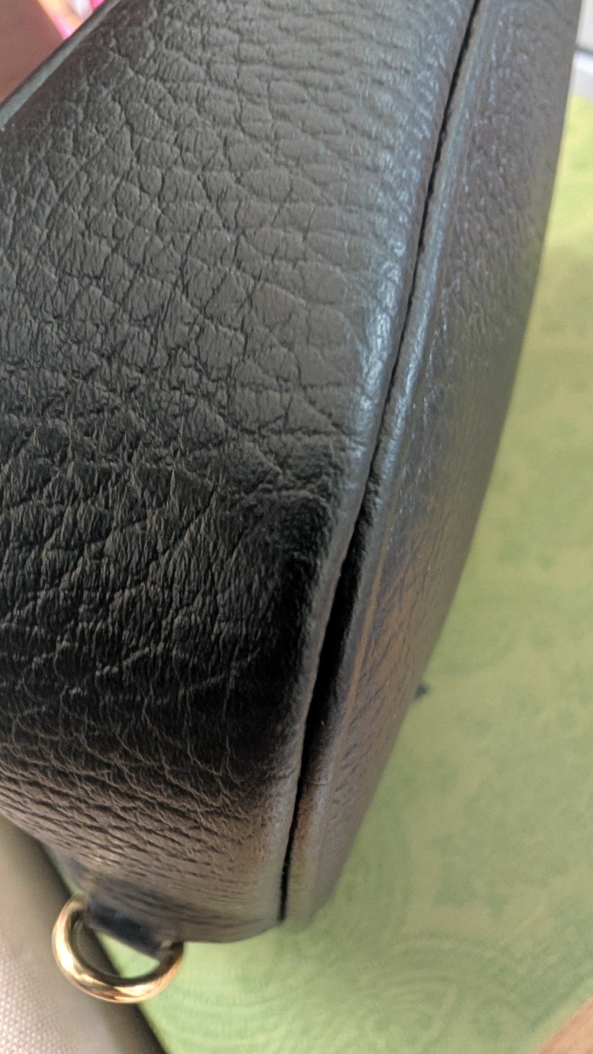 Close-up of a black pebbled leather bag corner showing a gold-tone metal ring attachment, with a green patterned surface in the background. Pre-Owned Gucci GG Marmont Black Leather Camera Crossbody Bag – Gold Hardware