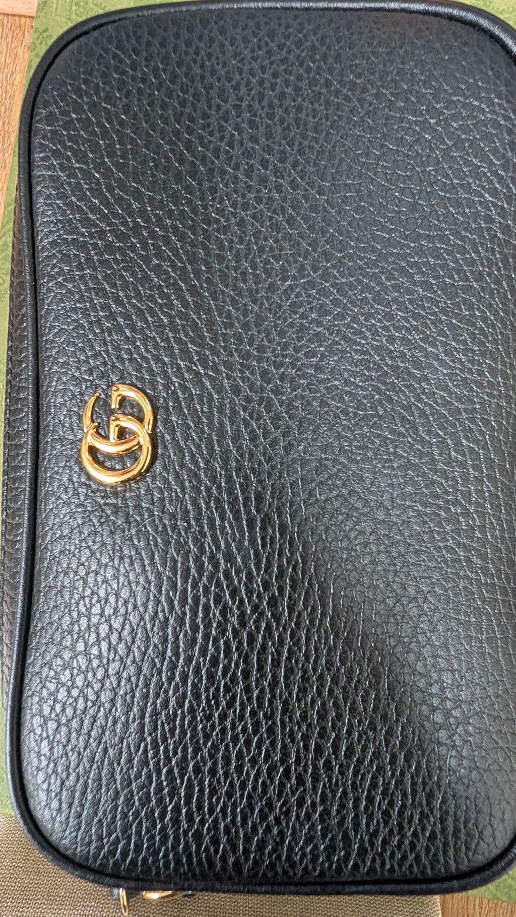 A close-up of a black pebbled leather pouch or small bag with a gold-tone interlocking double-G style logo on the front, photographed against a small glimpse of patterned fabric at the edges. Pre-Owned Gucci GG Marmont Black Leather Camera Crossbody Bag – Gold Hardware
