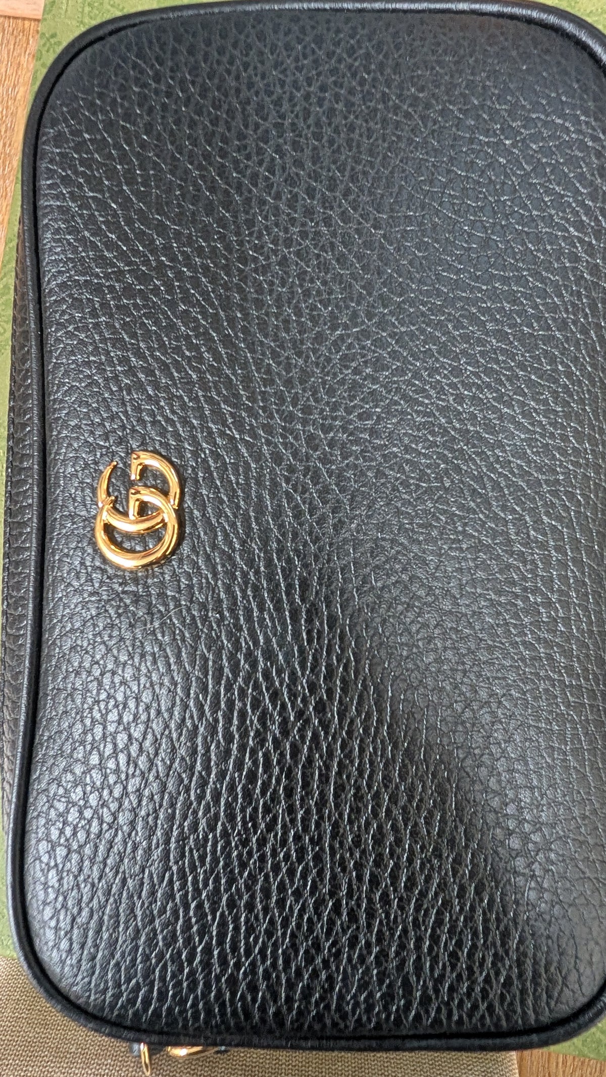 A close-up of a black pebbled leather pouch or small bag with a gold-tone interlocking double-G style logo on the front, photographed against a small glimpse of patterned fabric at the edges. Pre-Owned Gucci GG Marmont Black Leather Camera Crossbody Bag – Gold Hardware