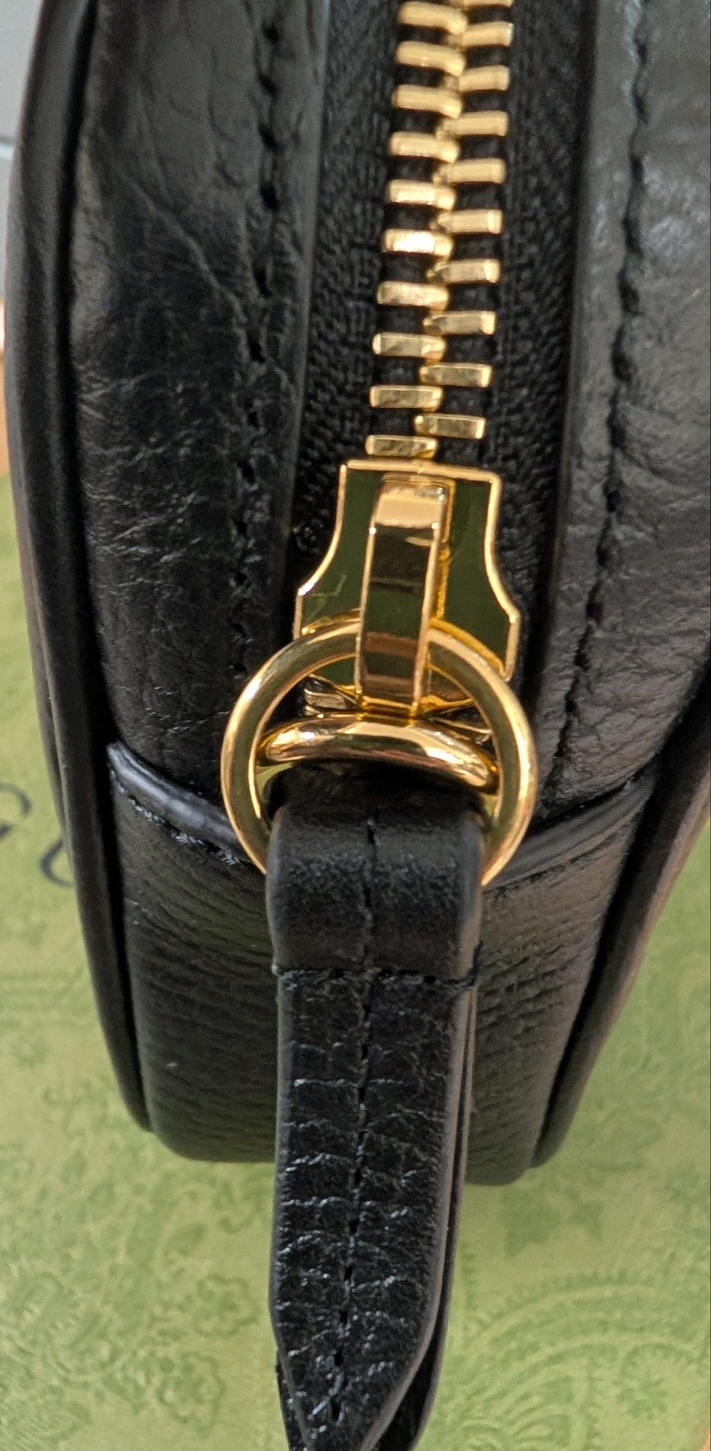 Close-up of a black leather handbag showing a gold-tone zipper pull and ring detail, resting on a green patterned surface. Pre-Owned Gucci GG Marmont Black Leather Camera Crossbody Bag – Gold Hardware