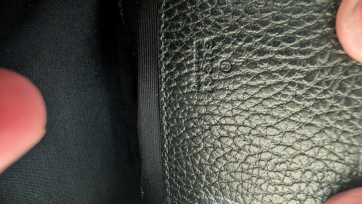 Close-up of a black textured leather interior panel with a visible 'GUCCI' logo stamp, next to black fabric lining, with fingers touching the edges. Pre-Owned Gucci GG Marmont Black Leather Camera Crossbody Bag – Gold Hardware
