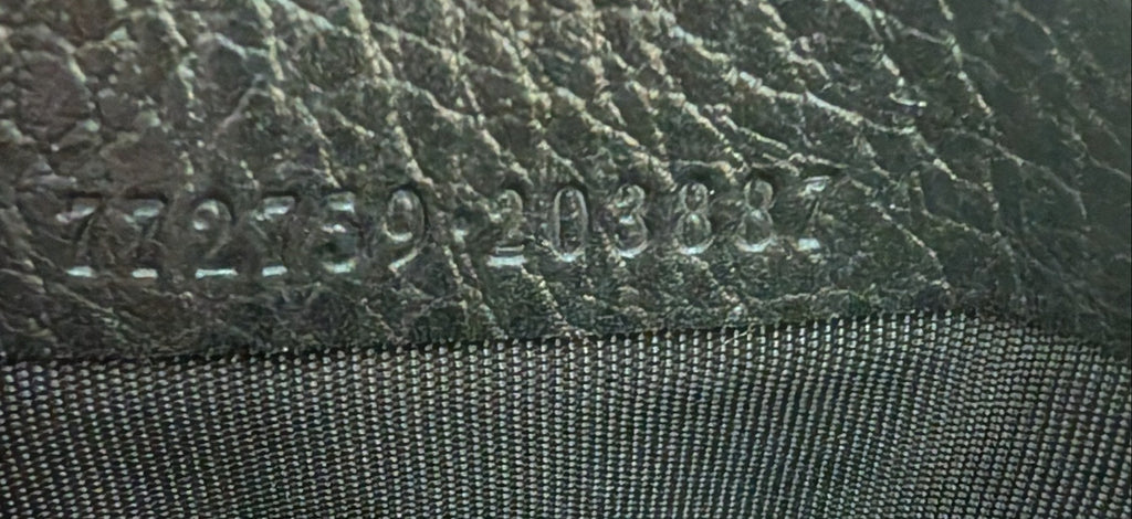 Close-up of a black leather 
 embossed serial numbers, held by fingers next to black textured fabric. Pre-Owned Gucci GG Marmont Black Leather Camera Crossbody Bag – Gold Hardware