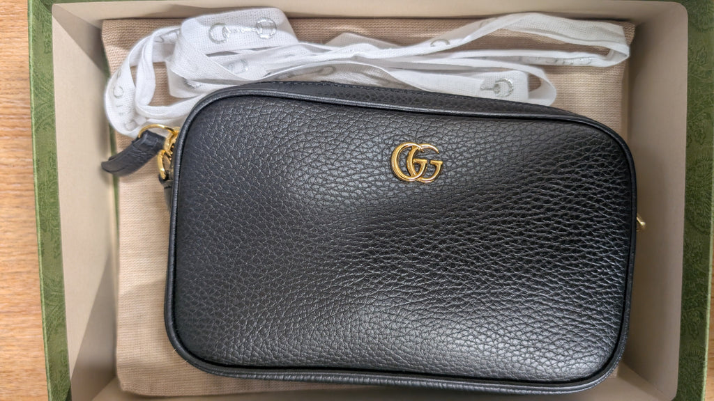 A black pebbled leather crossbody bag with a gold interlocking G logo, shown inside its original box with a white dust bag. Pre-Owned Gucci GG Marmont Black Leather Camera Crossbody Bag – Gold Hardware