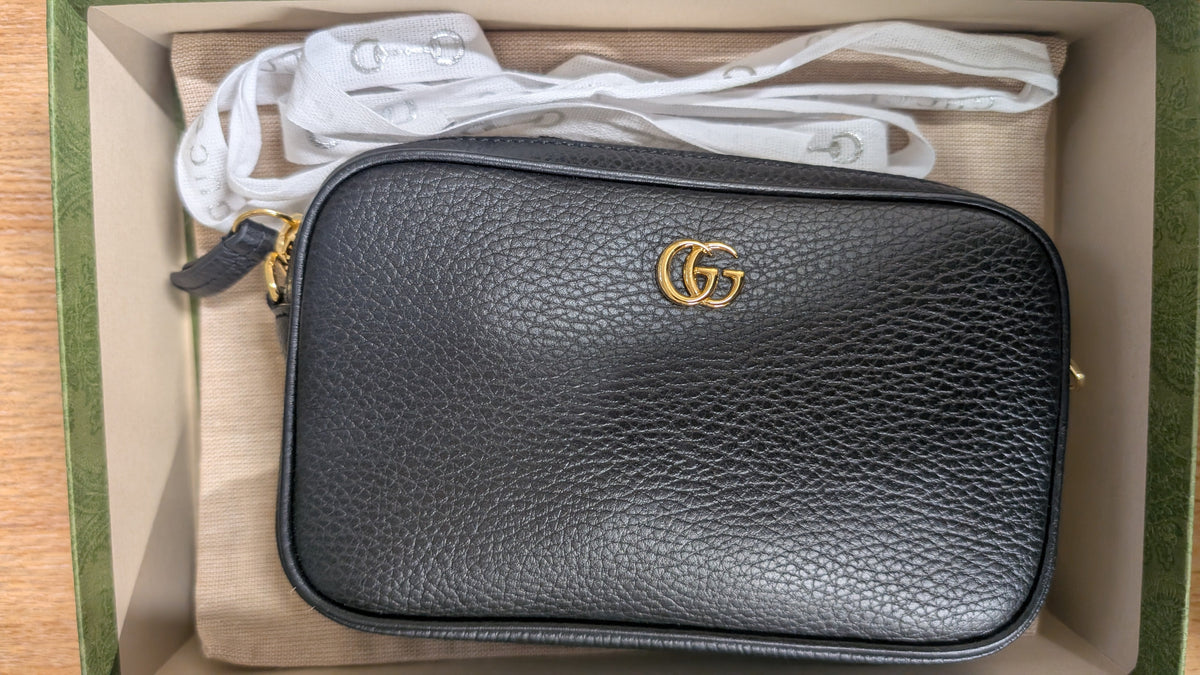 A black pebbled leather crossbody bag with a gold interlocking G logo, shown inside its original box with a white dust bag. Pre-Owned Gucci GG Marmont Black Leather Camera Crossbody Bag – Gold Hardware