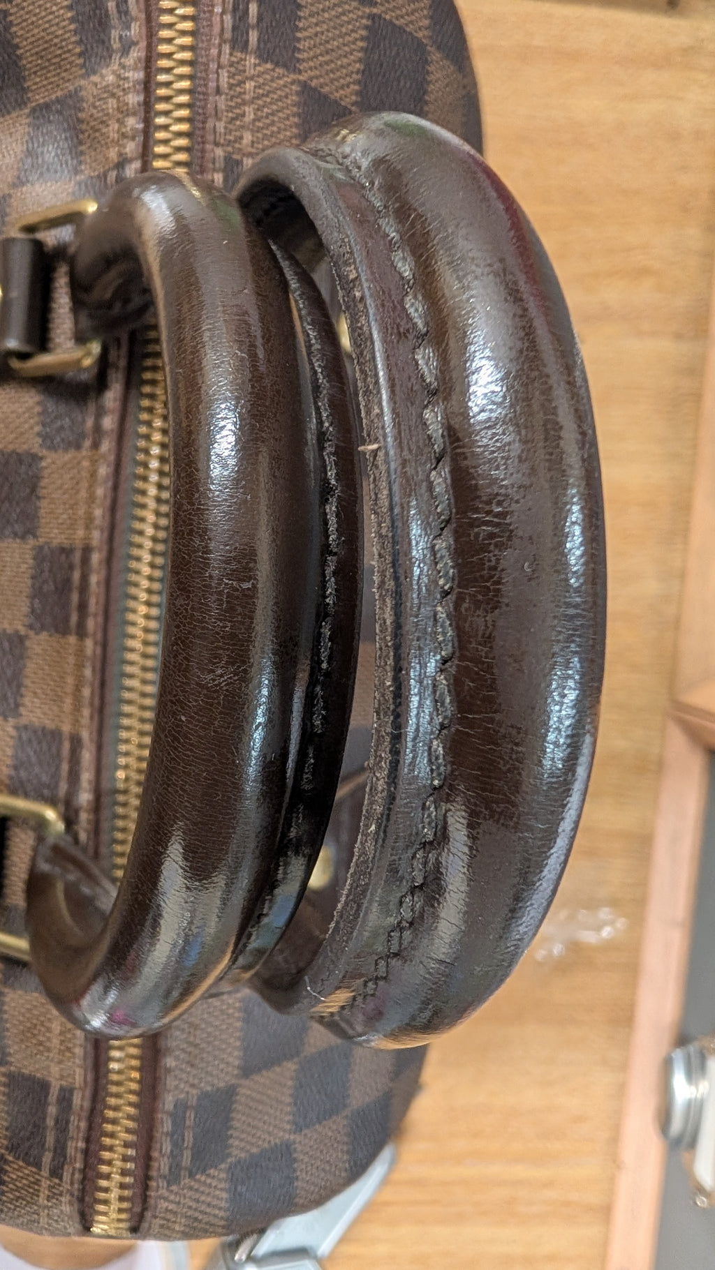 Close-up of a dark brown leather handle with visible wear and scuffs, attached to a brown and beige checkered canvas bag with gold-tone hardware. Pre-Owned Louis Vuitton Speedy 25 Damier Ebene Coated Canvas Bag DU4151