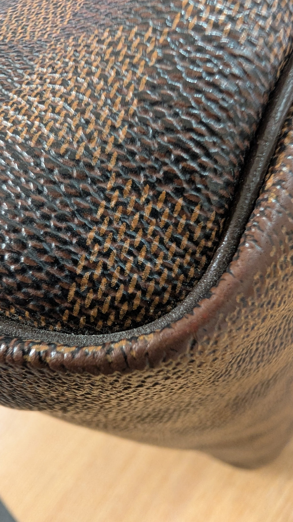 Close-up of a brown textured leather or coated canvas material with a woven pattern and stitched edge, shown against a light wood surface. Pre-Owned Louis Vuitton Speedy 25 Damier Ebene Coated Canvas Bag DU4151