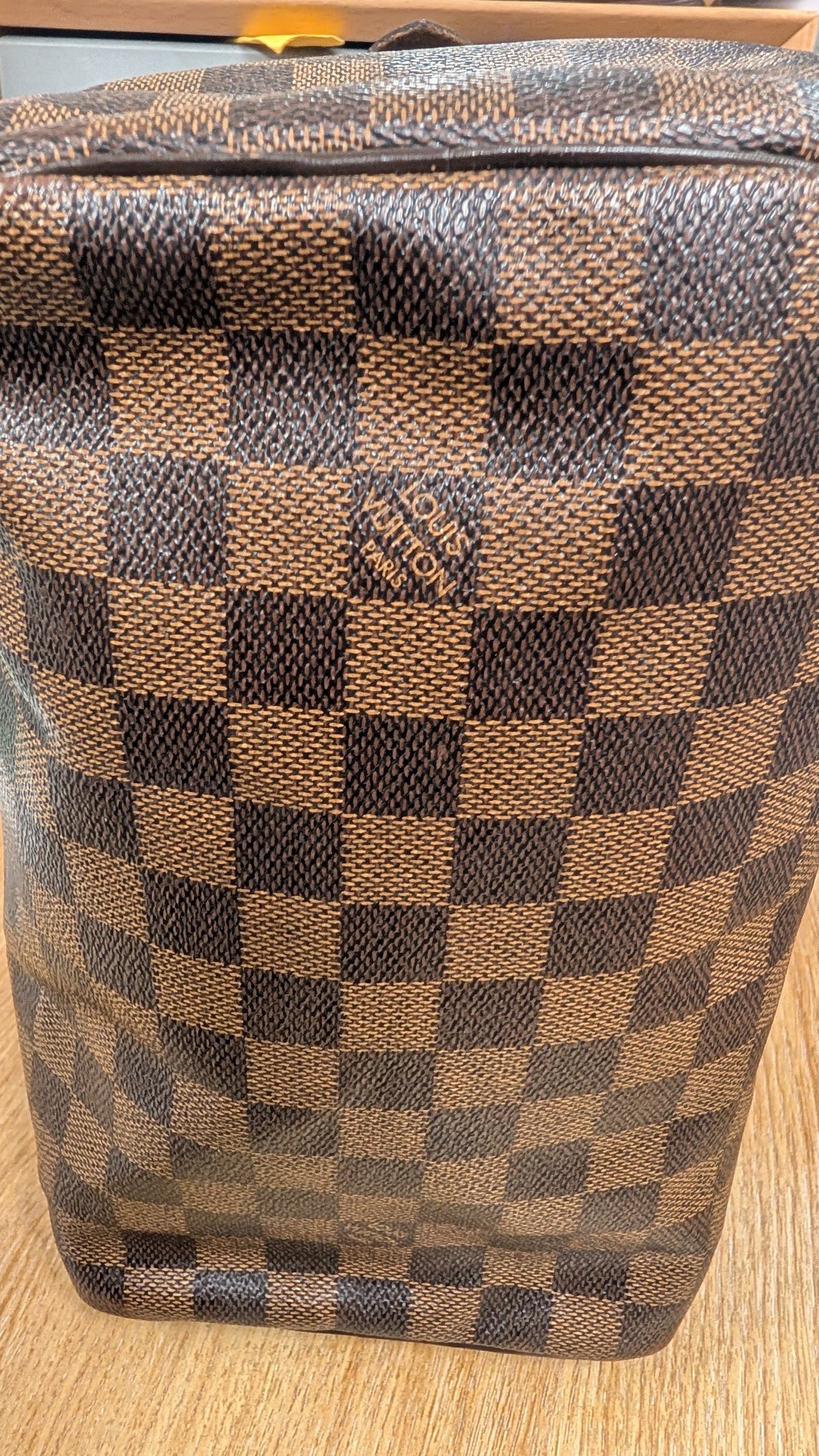 A Louis Vuitton  bag in the Damier Ebene checkered canvas, featuring a brown and beige pattern with a small gold-tone brand logo on the front, set on a light wood surface. Pre-Owned Louis Vuitton Speedy 25 Damier Ebene Coated Canvas Bag DU4151