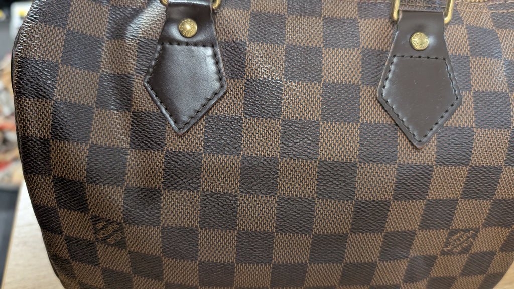 Close-up of a brown and dark brown checkerboard patterned handbag with dark brown leather strap attachments and gold-tone rivets. Pre-Owned Louis Vuitton Speedy 25 Damier Ebene Coated Canvas Bag DU4151