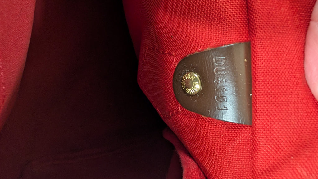 Close-up of a red canvas or fabric bag interior showing a brown leather patch with a gold-toned rivet and embossed lettering. Pre-Owned Louis Vuitton Speedy 25 Damier Ebene Coated Canvas Bag DU4151.