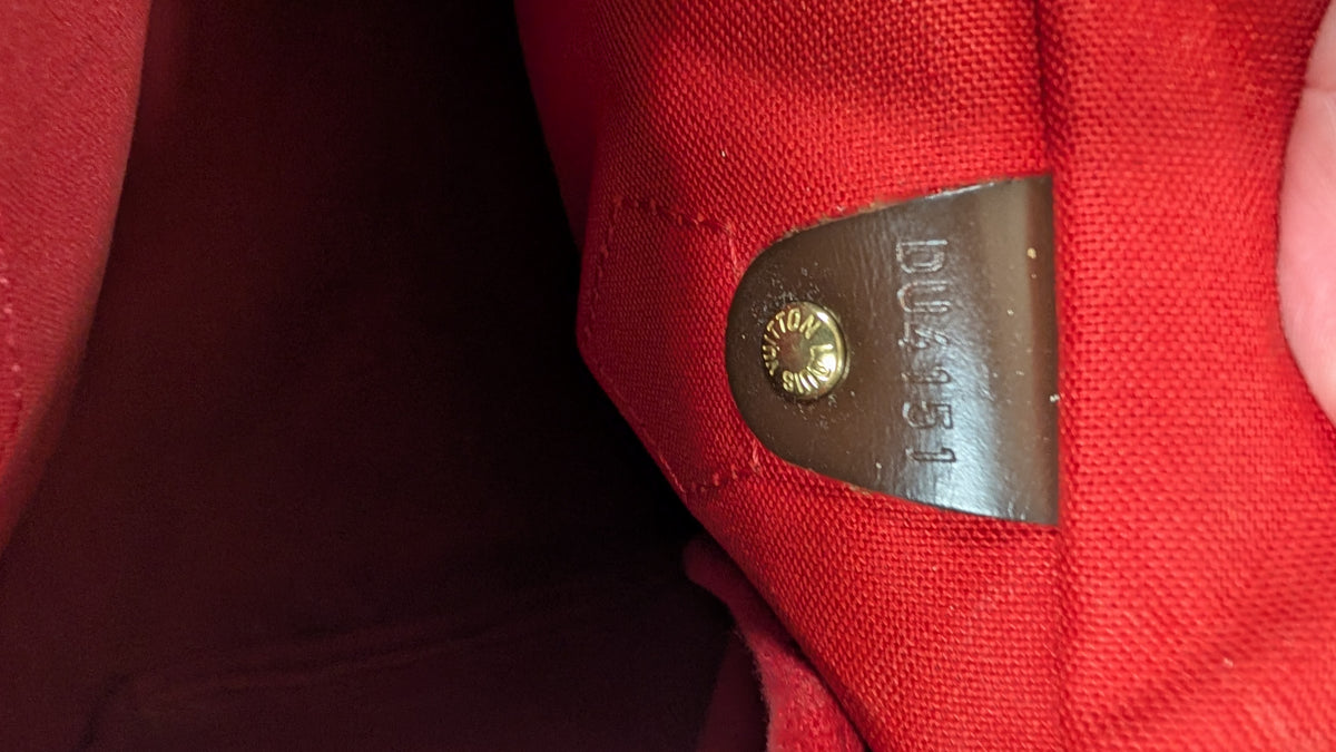 Close-up of a red canvas or fabric bag interior showing a brown leather patch with a gold-toned rivet and embossed lettering. Pre-Owned Louis Vuitton Speedy 25 Damier Ebene Coated Canvas Bag DU4151.