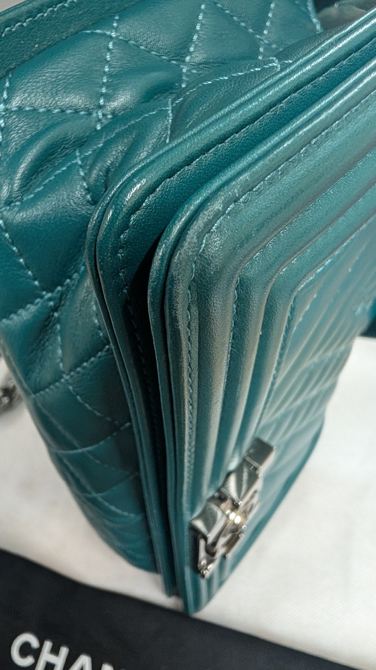 Close-up of a teal quilted leather handbag showing the texture, stitching, and silver-tone hardware, with a partial Chanel logo visible on the dust bag. 