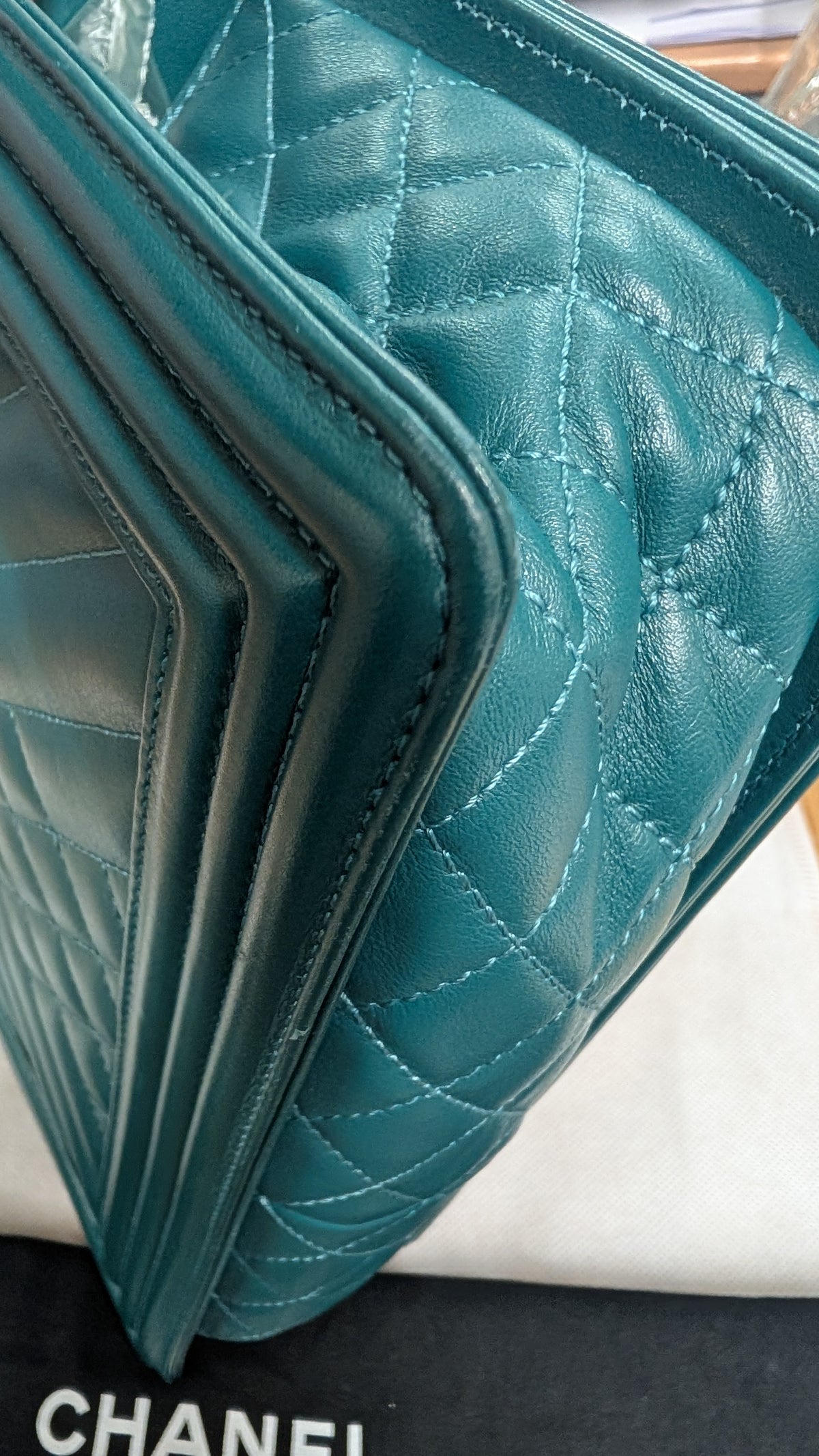 A close-up of a teal quilted leather handbag, showing the texture and stitching, with a black dust bag featuring the Chanel logo at the bottom.   Pre-Owned Chanel Boy Hang Bag – Teal Quilted Leather with Ruthenium-Tone Hardware