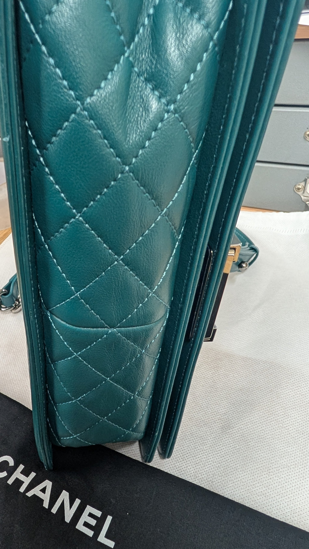 A close-up of a teal quilted leather handbag, showing the diamond-stitched pattern and side profile, resting on a white surface with a black Chanel dust bag visible in the foreground. Pre-Owned Chanel Boy Hang Bag – Teal Quilted Leather with Ruthenium-Tone Hardware