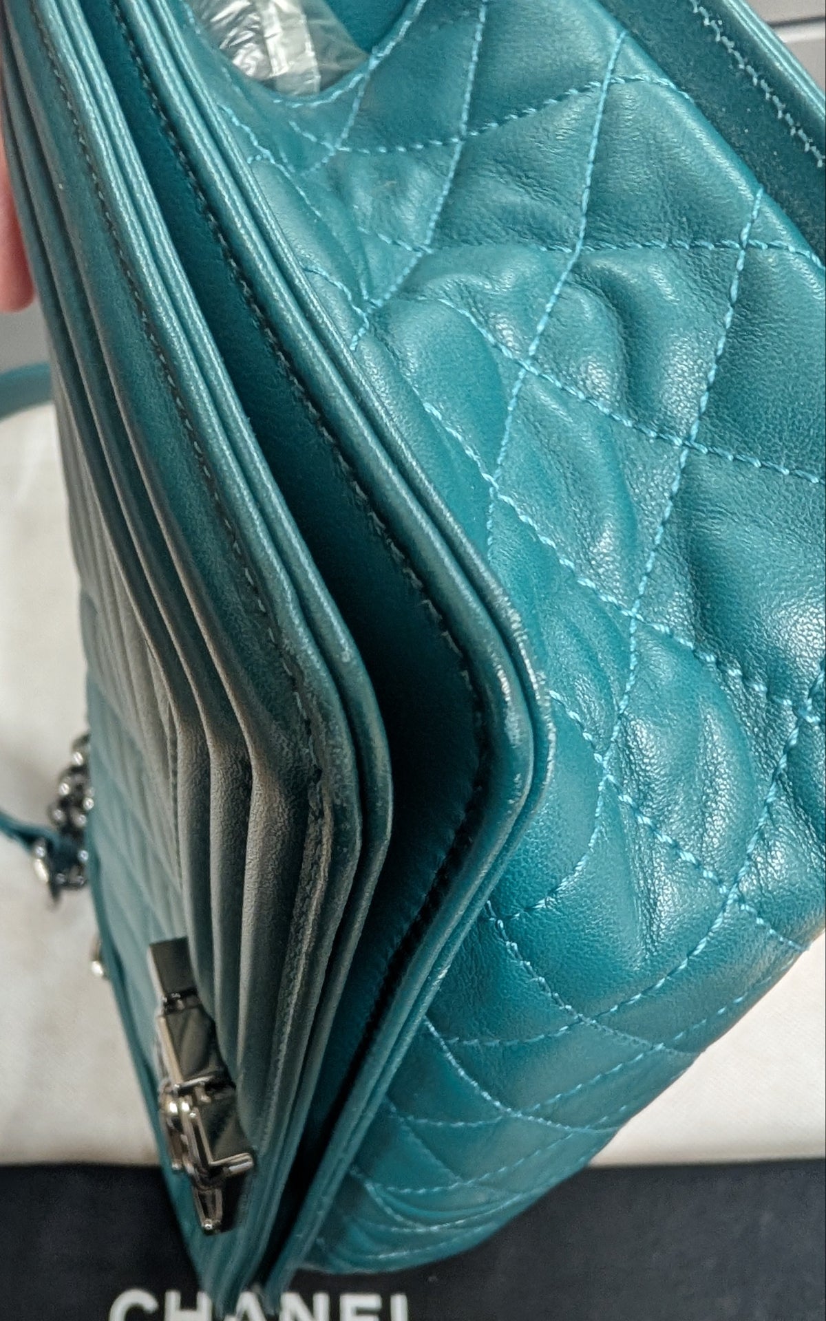 A close-up of a teal quilted Chanel shoulder bag with silver hardware, held next to a black Chanel dust bag on a light surface. Pre-Owned Chanel Boy Hang Bag – Teal Quilted Leather with Ruthenium-Tone Hardware