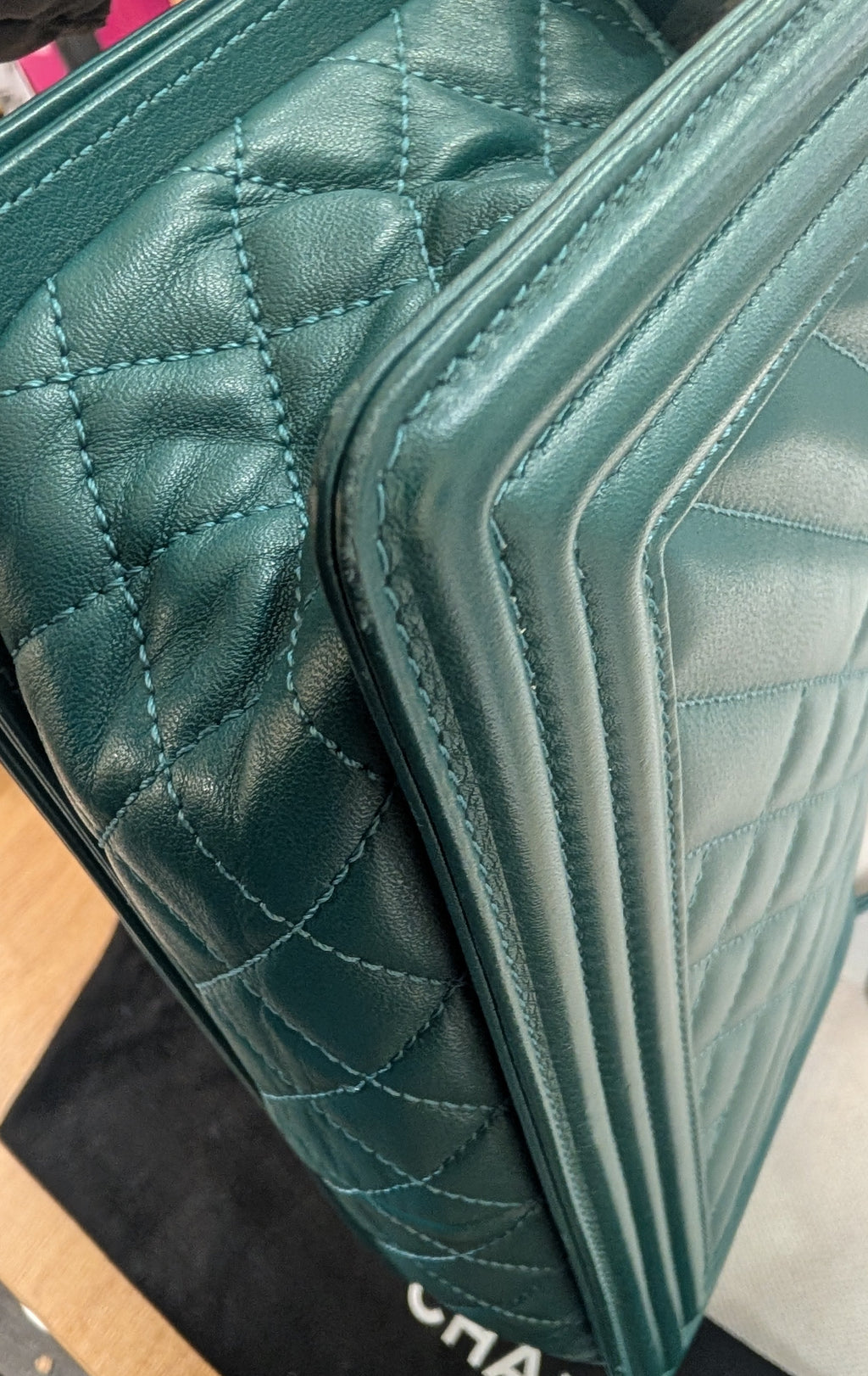 A close-up of a teal green quilted leather handbag with visible stitching and a fold-over flap, resting on a black Chanel dust bag. Pre-Owned Chanel Boy Hang Bag – Teal Quilted Leather with Ruthenium-Tone Hardware