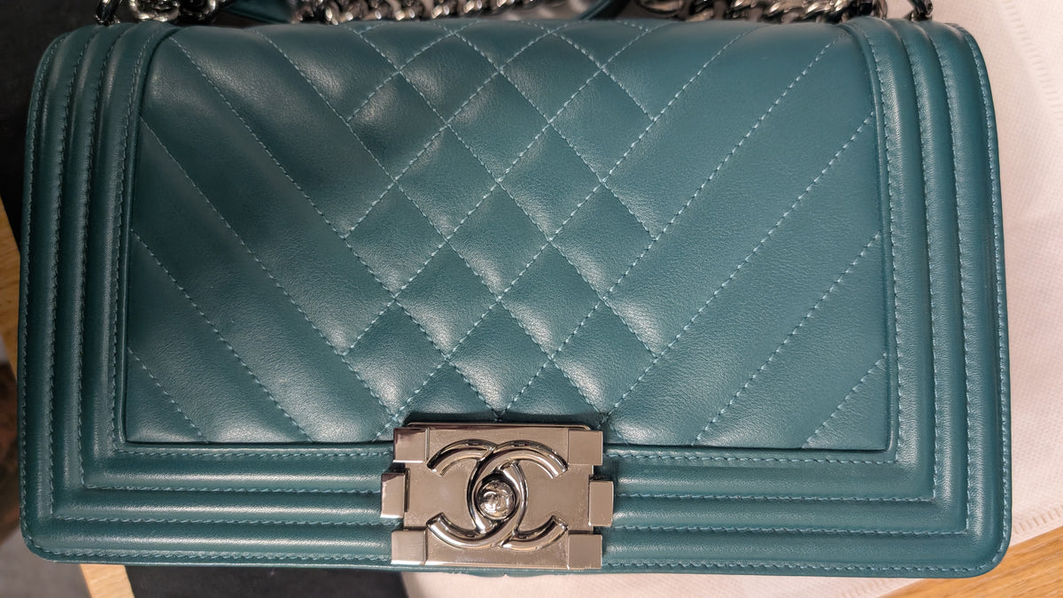 A quilted teal blue leather shoulder bag with a silver-tone interlocking CC clasp, characteristic of a Chanel Boy bag, shown on a light surface. Pre-Owned Chanel Boy Hang Bag – Teal Quilted Leather with Ruthenium-Tone Hardware