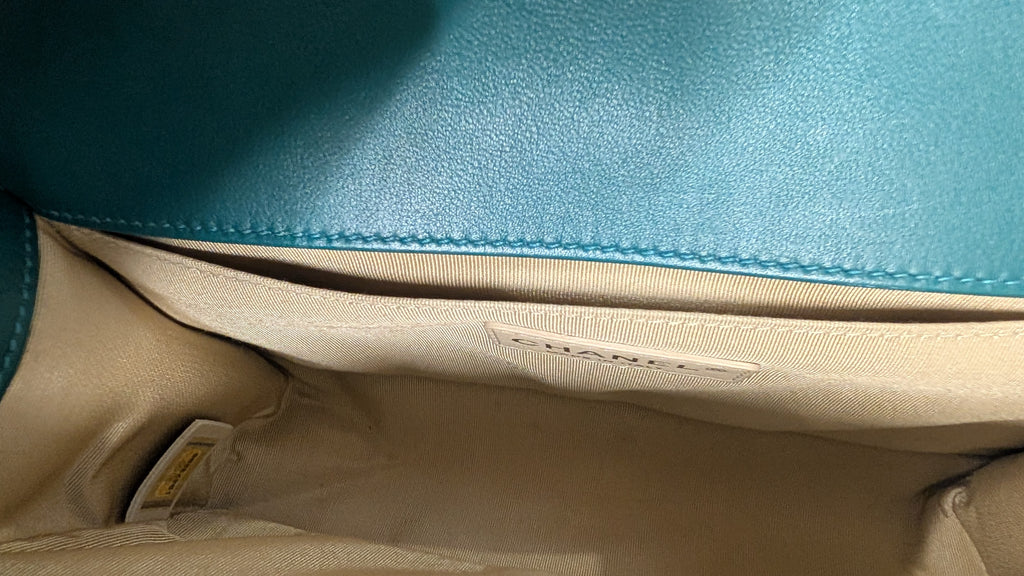 Close-up of the interior of a handbag, showing a teal blue leather exterior stitched to a beige fabric lining. A small fabric label is stitched on the lining, and a product tag is visible in the bottom left corner. Pre-Owned Chanel Boy Hang Bag – Teal Quilted Leather with Ruthenium-Tone Hardware