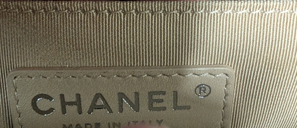 Close-up of a Chanel interior label sewn onto a beige fabric strap, with fingers holding the strap open to show the stitching and brand text. A turquoise leather-like surface is visible at the right edge. Pre-Owned Chanel Boy Hang Bag – Teal Quilted Leather with Ruthenium-Tone Hardware