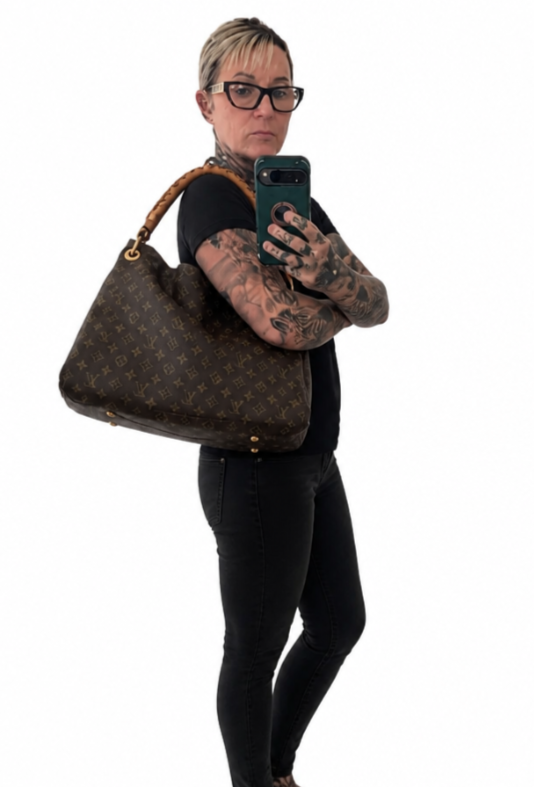 A person taking a mirror selfie while wearing a large brown Louis Vuitton monogram shoulder bag with a tan leather handle. The person is wearing a black t-shirt, black pants, and glasses, and has visible tattoos on their arms. Pre-Owned Louis Vuitton Artsy MM Monogram Canvas Shoulder Bag 